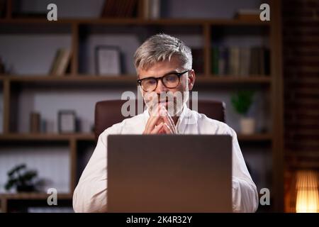 Gray-haired manager in glasses uses a laptop, conducts a video conference in the office. Middle aged businessman participating in online webinar Stock Photo