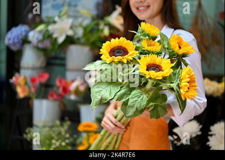 Charming and beautiful Asian female florist or flower shop worker holding a beautiful sunflower bouquet. cropped and close-up image Stock Photo