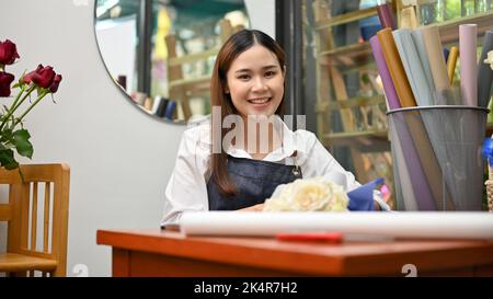 Cheerful and smiling young Asian female florist or floral shop arranging a beautiful white roses bouquet in her shop. Small business startup concept Stock Photo