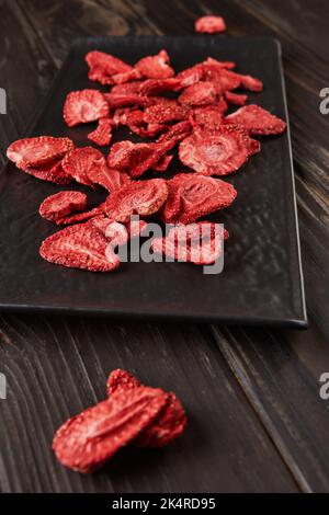 Strawberry chips are stacked in black ceramic tray with dark wooden background. Hundred-percent natural freeze-dried strawberries Stock Photo