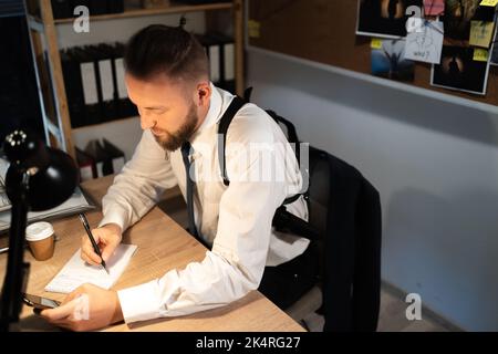 professional detective works on a murder in the office at night. He sits at the table, studying the evidence, making notes in his notebook. Stock Photo