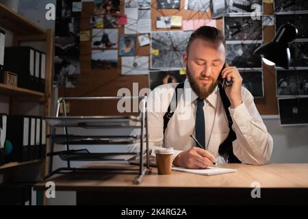 A professional detective works on a murder in the office at night. Sitting at the table takes a call from a witness and writes down the testimony in a Stock Photo