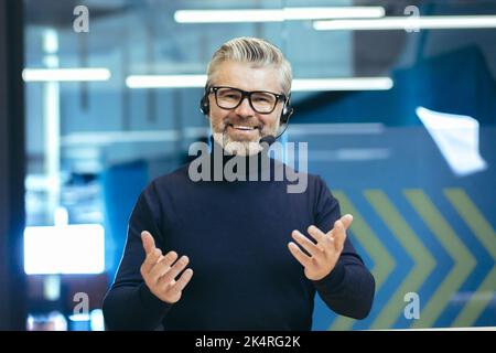 Online business training. A young handsome gray-haired man in headphones with a microphone conducts an online meeting, webinar on camera. He waves his hands, smiles. Stock Photo
