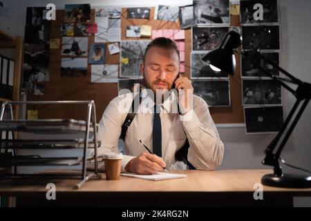 A professional detective works on a murder in the office at night. Sitting at the table takes a call from a witness and writes down the testimony in a Stock Photo