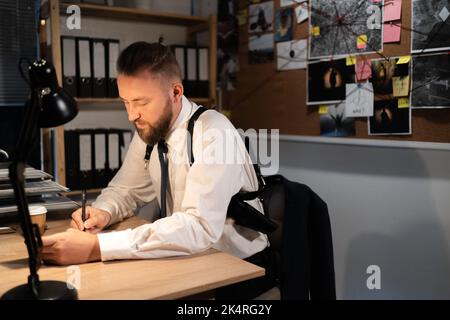 A professional detective works on a murder in the office at night. He sits at the table, studying the evidence, making notes in notebook. Stock Photo