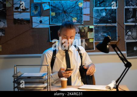 A professional detective works on a murder in the office at night. He sits at the table, holds a smartphone in his hands and reads the news online. Stock Photo
