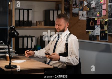 A professional detective works on a murder in the office at night. He sits at the table, holds a smartphone in his hands and reads the news online. Stock Photo