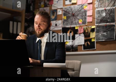 Detective working in office using computer at night, drinking coffee at workplace Stock Photo