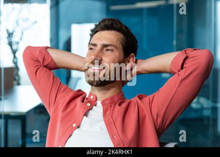 Young successful businessman startuper rests in office, man in red shirt throws hands behind head and dreams visualizes, designer thinks about new project. Stock Photo
