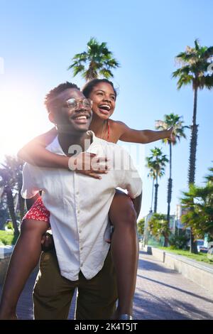 Cheerful vertical portrait of a young mixed race couple doing piggyback on holidays on an island. Stock Photo