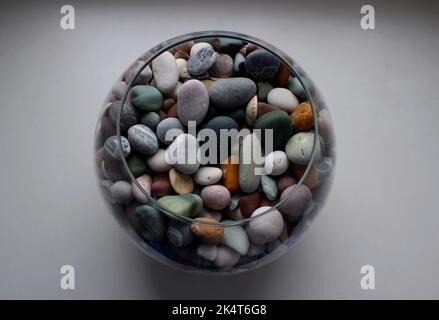 Round glass vase with colorful smooth pebbles and shells on a white surface isolated top view Stock Photo