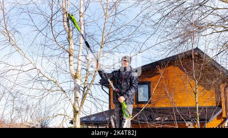 Electric chain pruner with a telescopic pole close up. Winter tree pruning. A man gardener standing on a stepladder cuts branches on a tree with a spe Stock Photo