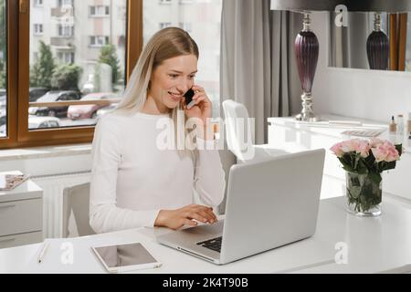 Woman works at the reception in a beauty salon. Typing something on laptop and talks on the phone with client, making appointments in studio Stock Photo