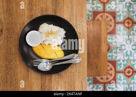 Fresh mango with sticky rice and sweetened coconut milk on wooden table. Popular Thai food prepared on plate to eat. Stock Photo