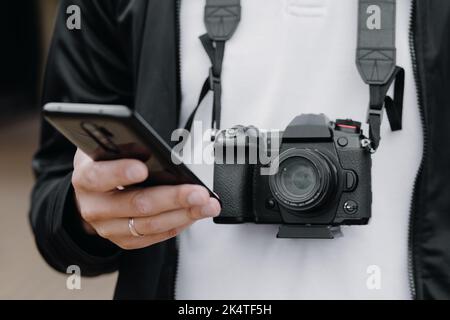 Photo of a European man holding a phone in his hands and a camera around his neck while walking outdoors. Look at the phone. Stock Photo
