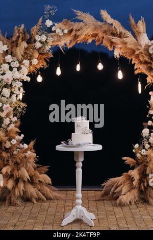 A white cake on a stand against the background of an evening wedding arch Stock Photo