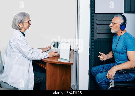 Audiological exam. Audiologist doing hearing check-up to senior man in soundproof audiometric booth. Hearing loss treatment, side view Stock Photo
