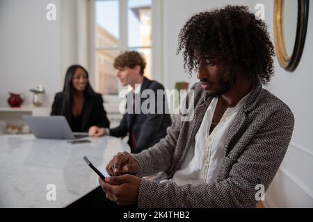 African businessman uses his smartphone in the office while his colleagues discuss work Stock Photo