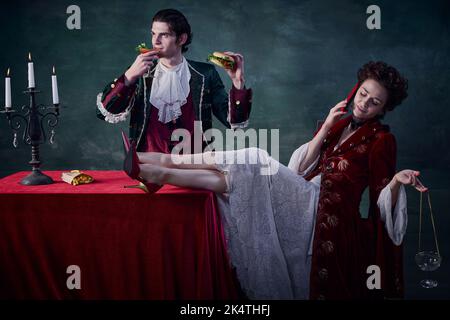 Portrait of man and woman in image of medieval vampires over dark green background. Man eating burger and drinking bloody mary, woman talking on phone Stock Photo