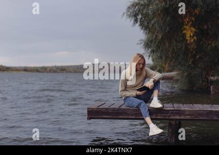 In autumn, a girl on the riverbank reads a book Stock Photo