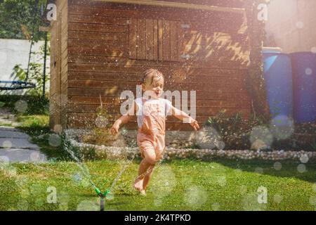 Girl in wet clothes run around water sprinkler at garden Stock Photo