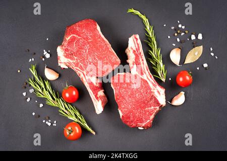 Raw ribeye beef steaks with seasonings over dark concrete background. Cowboy steak on bone ready for grilling. View above Stock Photo