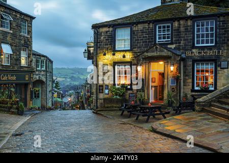 Haworth Main Street (steep hill, old buildings, blue hour evening light, historic Bronte sisters' village, Grade 2 pub) - West Yorkshire, England, UK. Stock Photo