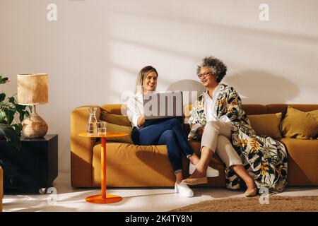Two happy businesswomen using a laptop while working together in an office lobby. Successful female entrepreneurs sitting on a couch in a woman-owned Stock Photo