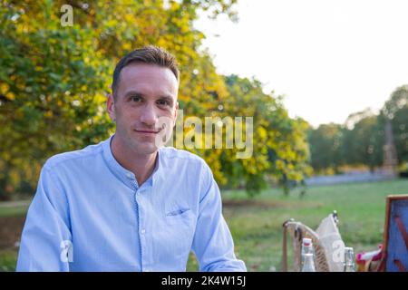 Young man in his 30s sitting on picnic mat at park smiles looking straight ahead, copy space on the right Stock Photo