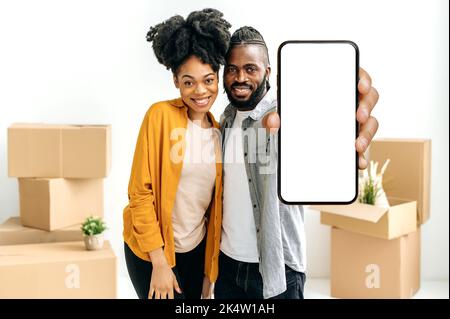 Happy married african american couple, newlyweds, show smart phone with empty white mock-up screen while stand in living room against the background of cardboard boxes with things in their new home Stock Photo