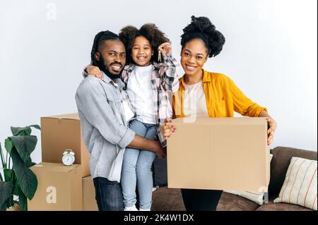 New housing, relocation. Happy african american family, dad mom and their cute daughter, moved to their new apartment, holding boxes with things, girl in dad's arms shows keys, they smile into camera Stock Photo
