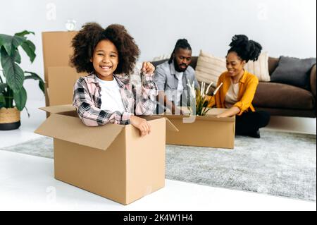 An African American family moves into their new home. Happy cute girl preschooler, sits in a box in the living room, shows the keys, looks at camera, smile, parents are sorting out a box in background Stock Photo