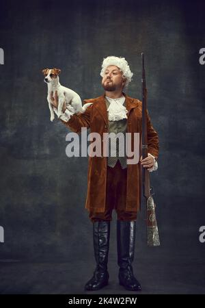 Companion. Portrait of young man in image of nobleman in vintage brown hunting suit and white wig holding cute little dog isolated over dark retro Stock Photo