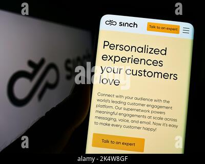 Person holding cellphone with website of Swedish communications company Sinch AB on screen in front of logo. Focus on center of phone display. Stock Photo