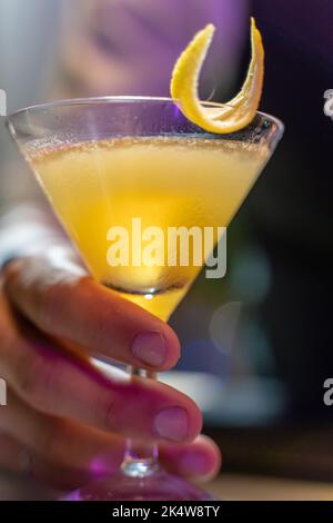 Close-up of a barman's hand serving a beautiful yellow cocktail with lemon peel on the bar counter in the nightclub. Stock Photo