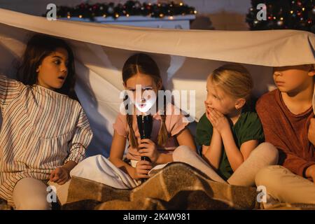 Girl holding flashlight near scared friends under blanket during christmas at home,stock image Stock Photo