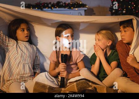 Girl holding flashlight near scared interracial friends under blanket and christmas decor at home,stock image Stock Photo