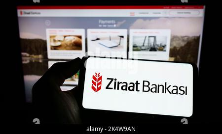 Person holding cellphone with logo of Turkish financial services company Ziraat Bank on screen in front of webpage. Focus on phone display. Stock Photo