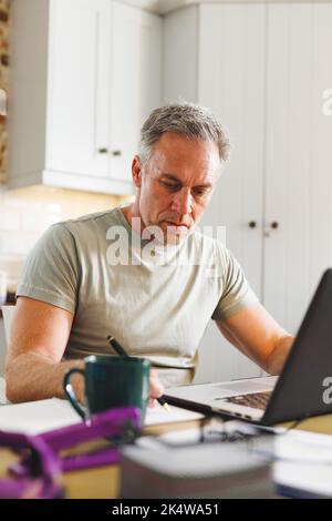 Happy caucasian man sitting at table in kitchen and using laptop and taking notes Stock Photo