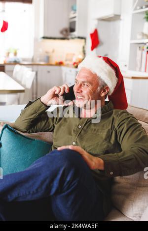 Happy caucasian man wearing santa hat, sitting on sofa in living room, talking on smartphone Stock Photo