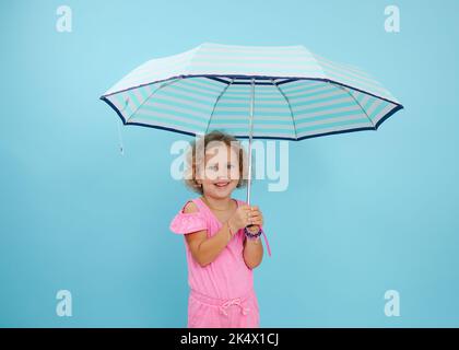 Portrait of little girl with curly fair hair wearing pink jumpsuit, holding open striped umbrella on blue background. Stock Photo