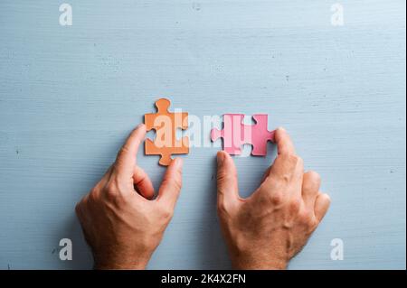Top view of male hands holding two matching puzzle pieces in orange and pink colores. Over blue wooden background. Stock Photo