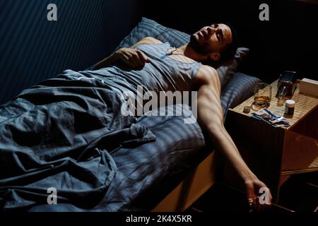 Young stressed man trying to get asleep while lying on bed by night table with whisky in bottle and glass, sleeping pills and photos Stock Photo