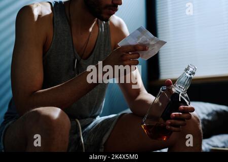 Young depressed man with alcoholic drink looking at picture of him on bike while suffering from post traumatic syndrome and insomnia Stock Photo