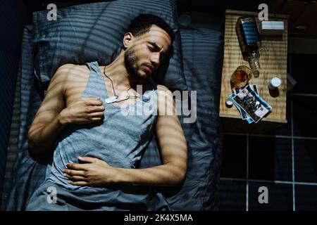 Young man with his eyes closed having nightmare or trying to get asleep while lying in bed by night table with alcohol, photos and pills Stock Photo