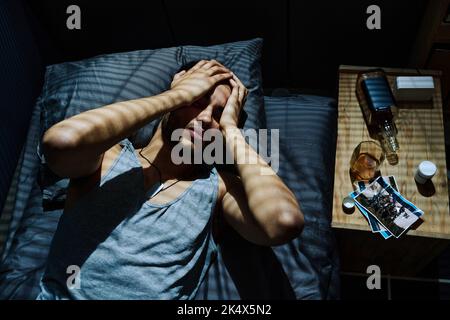 Young man suffering from headache caused by post traumatic syndrome or disorder while lying in bed and holding head in hands Stock Photo