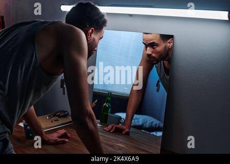 Young man with post traumatic disorder bending over table and looking at his reflection in mirror while suffering from insomnia Stock Photo