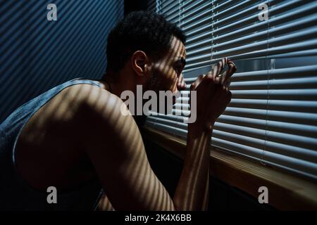 Side view view of young sleepless man looking through venetian blinds on window during sneak peek after someone outdoors Stock Photo