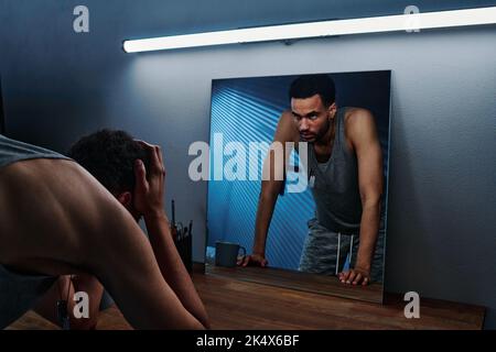 Young insomniac man holding head in hands in front of mirror with reflection of him expressing anger and aggression Stock Photo