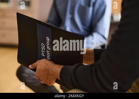 Black plastic folder in hands of young man with post traumatic syndrome sitting in front of psychotherapist and describing his problems Stock Photo